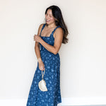 A woman wearing a blue floral print maxi dress and holding a small cream-colored handbag, posing against a plain white wall.
