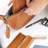 A person's hands are shown opening a tan leather zippered pouch to reveal its interior. A matching tan leather wallet and a blue textured phone case are laid out on a white table alongside other small accessories.
