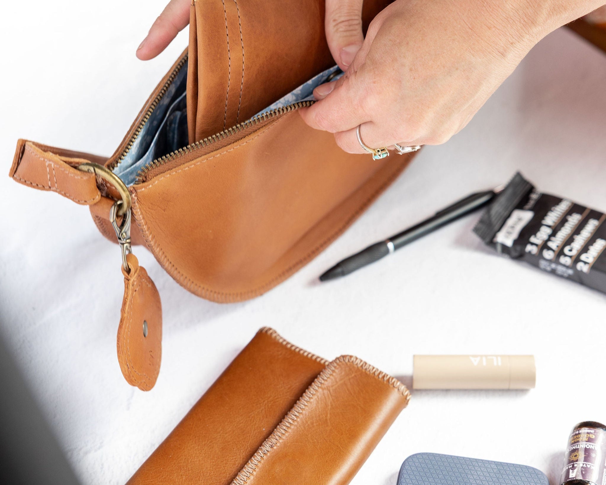 A person's hands are shown opening a tan leather zippered pouch to reveal its interior. A matching tan leather wallet and a blue textured phone case are laid out on a white table alongside other small accessories.