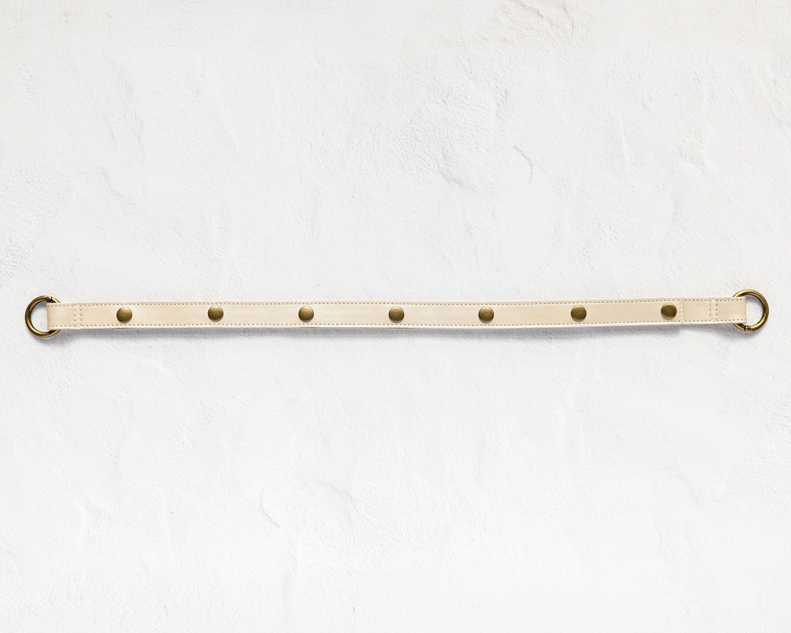 A beige leather belt with brass hardware, featuring five evenly spaced brass rivets and a matching brass ring on each end, laid horizontally on a textured white background.