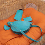 A turquoise blue leather shoulder bag with matching cardholder, wallet puffy heart charm, displayed on an orange cushion inside a wicker chair on a wooden deck.