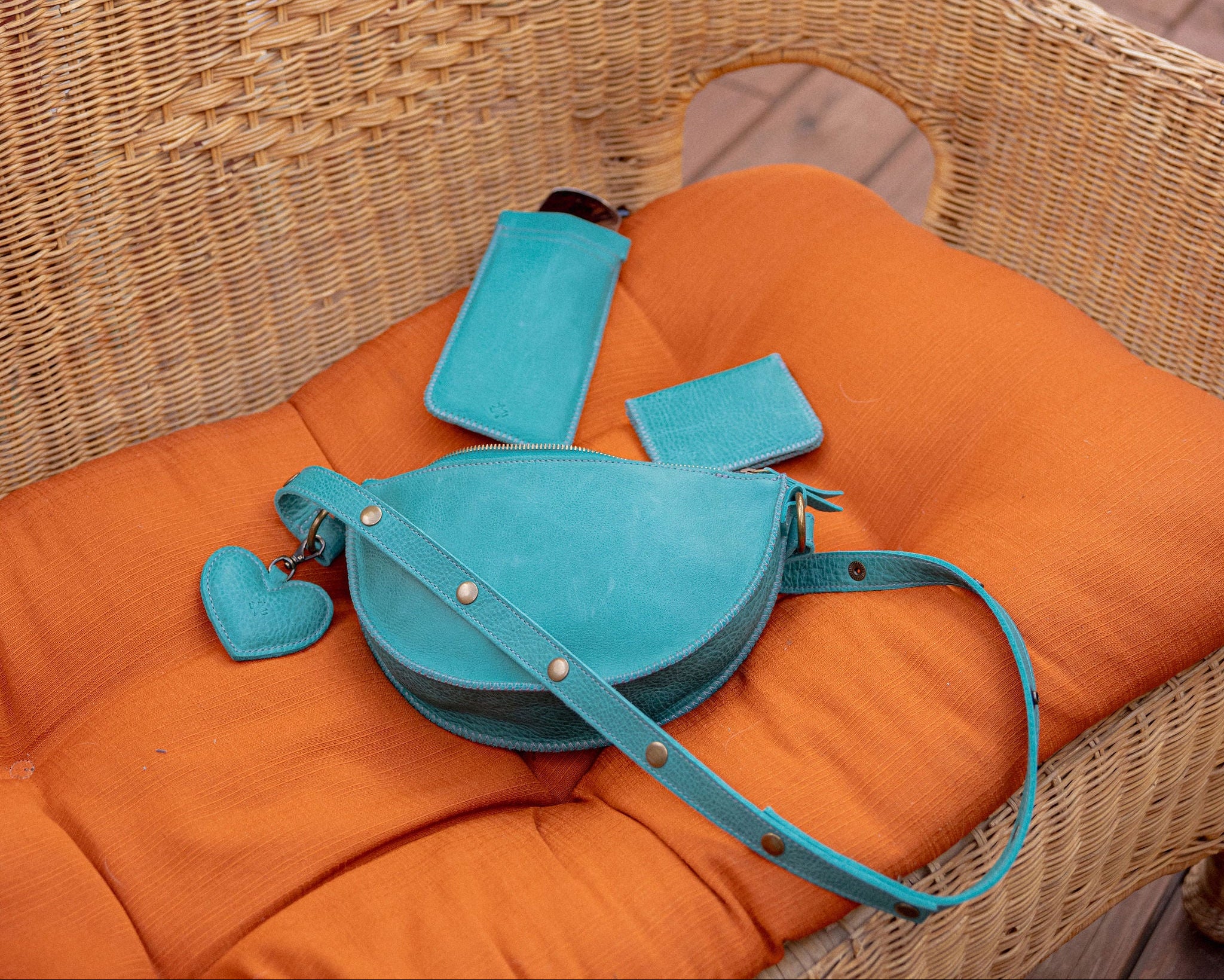 A turquoise blue leather shoulder bag with matching cardholder, wallet puffy heart charm, displayed on an orange cushion inside a wicker chair on a wooden deck.
