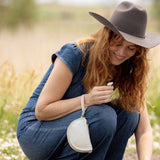 A woman crouching in a field of small white flowers, wearing a wide-brimmed brown hat, a blue denim jumpsuit, and holding a small white handbag.