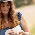 A woman outdoors wearing a brown cowboy hat and a blue denim overall dress, holding a white ceramic bowl with a small white flower in her hands.