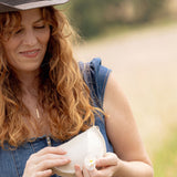 A woman outdoors wearing a brown cowboy hat and a blue denim overall dress, holding a white ceramic bowl with a small white flower in her hands.