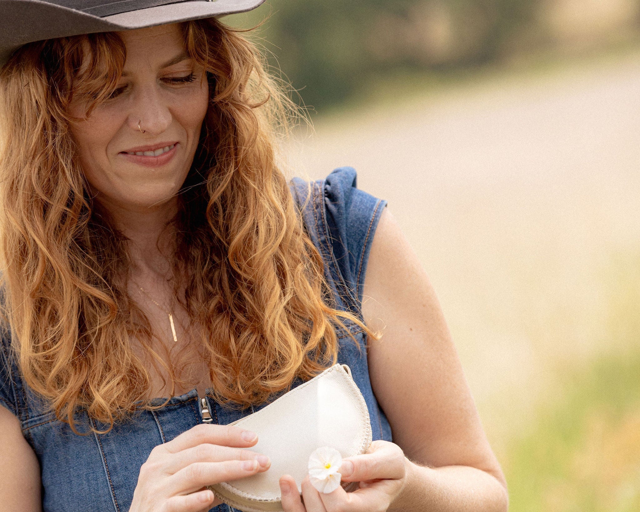 A woman outdoors wearing a brown cowboy hat and a blue denim overall dress, holding a white ceramic bowl with a small white flower in her hands.