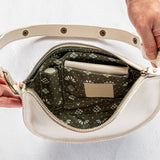 An open cream-colored leather shoulder bag with a patterned green and white floral lining, held open by two hands against a white textured background.