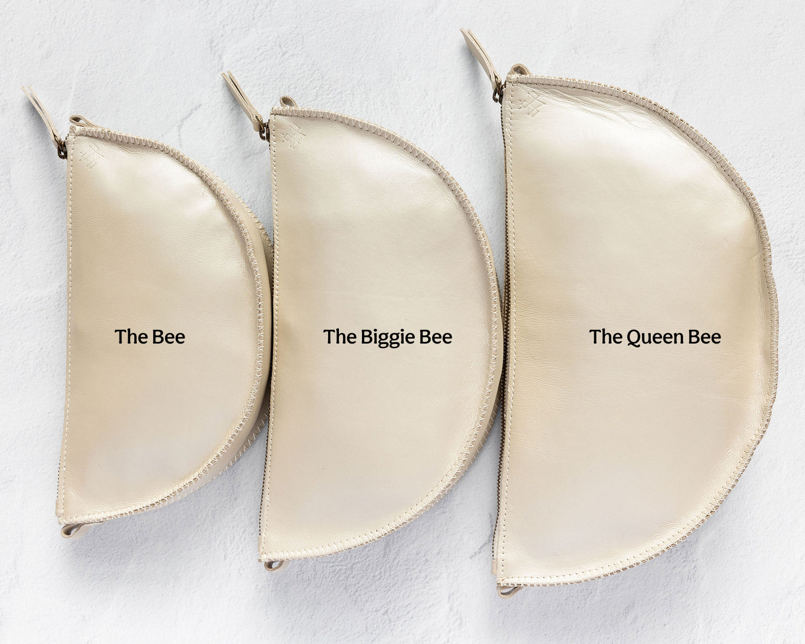 Three cream-colored leather half-moon zipper pouches of varying sizes arranged in a row on a textured white background, labeled 'The Bee', 'The Biggie Bee', and 'The Queen Bee'.