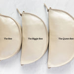 Three cream-colored leather half-moon zipper pouches of varying sizes arranged in a row on a textured white background, labeled 'The Bee', 'The Biggie Bee', and 'The Queen Bee'.