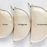 Three cream-colored leather half-moon zipper pouches of varying sizes arranged in a row on a textured white background, labeled 'The Bee', 'The Biggie Bee', and 'The Queen Bee'.