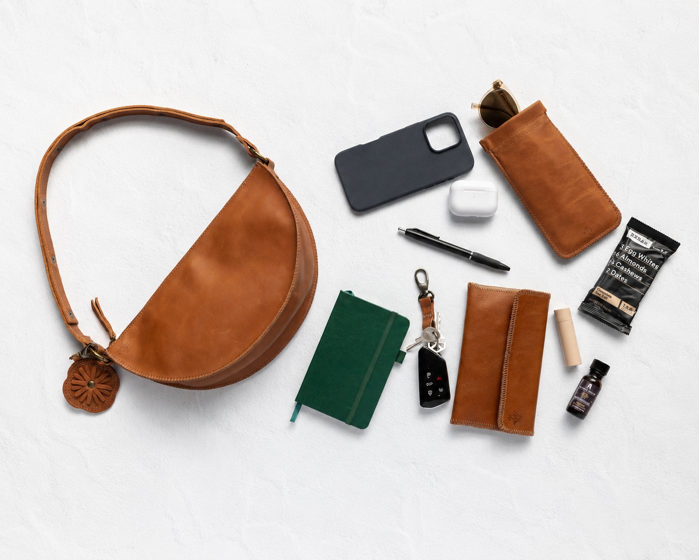 A styled flat-lay of a brown leather crescent shoulder bag on a white textured background, surrounded by various accessories including a green notebook, a black phone case, a pen, earbuds case, sunglasses, and a small bottle.