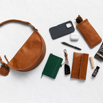 A styled flat-lay of a brown leather crescent shoulder bag on a white textured background, surrounded by various accessories including a green notebook, a black phone case, a pen, earbuds case, sunglasses, and a small bottle.