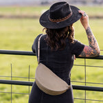 Person wearing a black hat and carrying a beige bag, standing in a field.