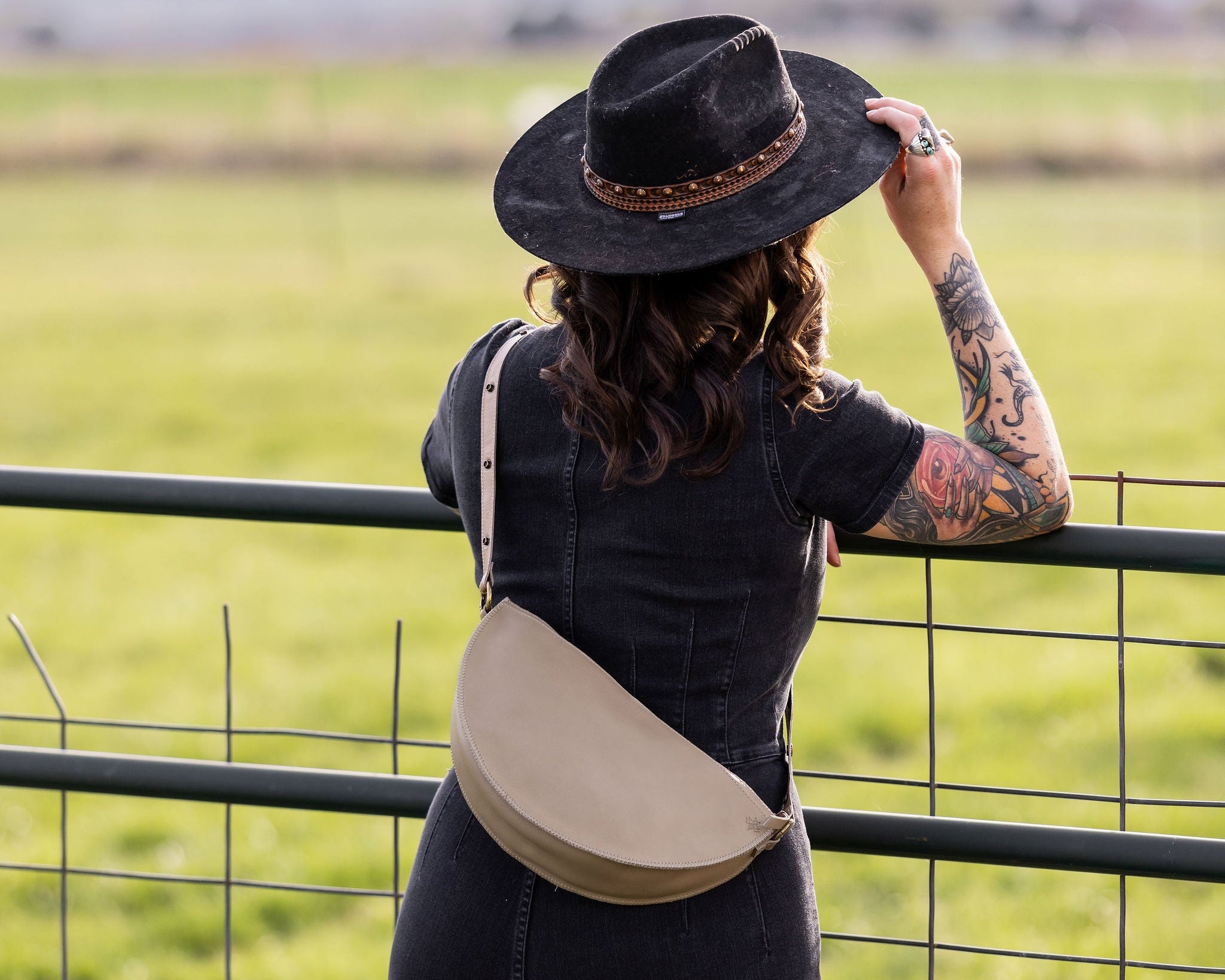 Person wearing a black hat and carrying a beige bag, standing in a field.