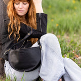 Woman sitting against a tree in a grassy field wearing a black jacket and light blue jeans.