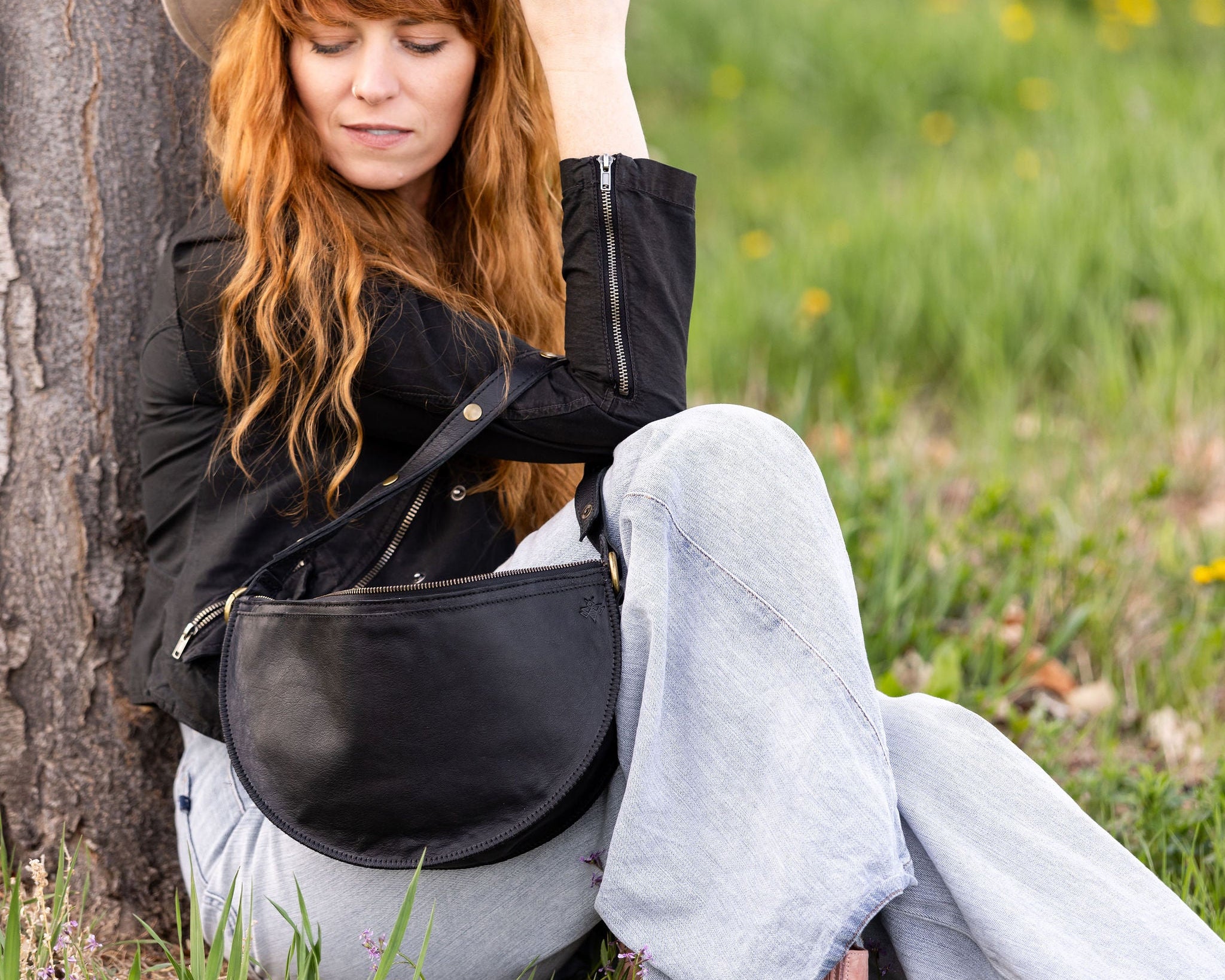 Woman sitting against a tree in a grassy field wearing a black jacket and light blue jeans.