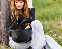 Woman sitting against a tree in a grassy field wearing a black jacket and light blue jeans.