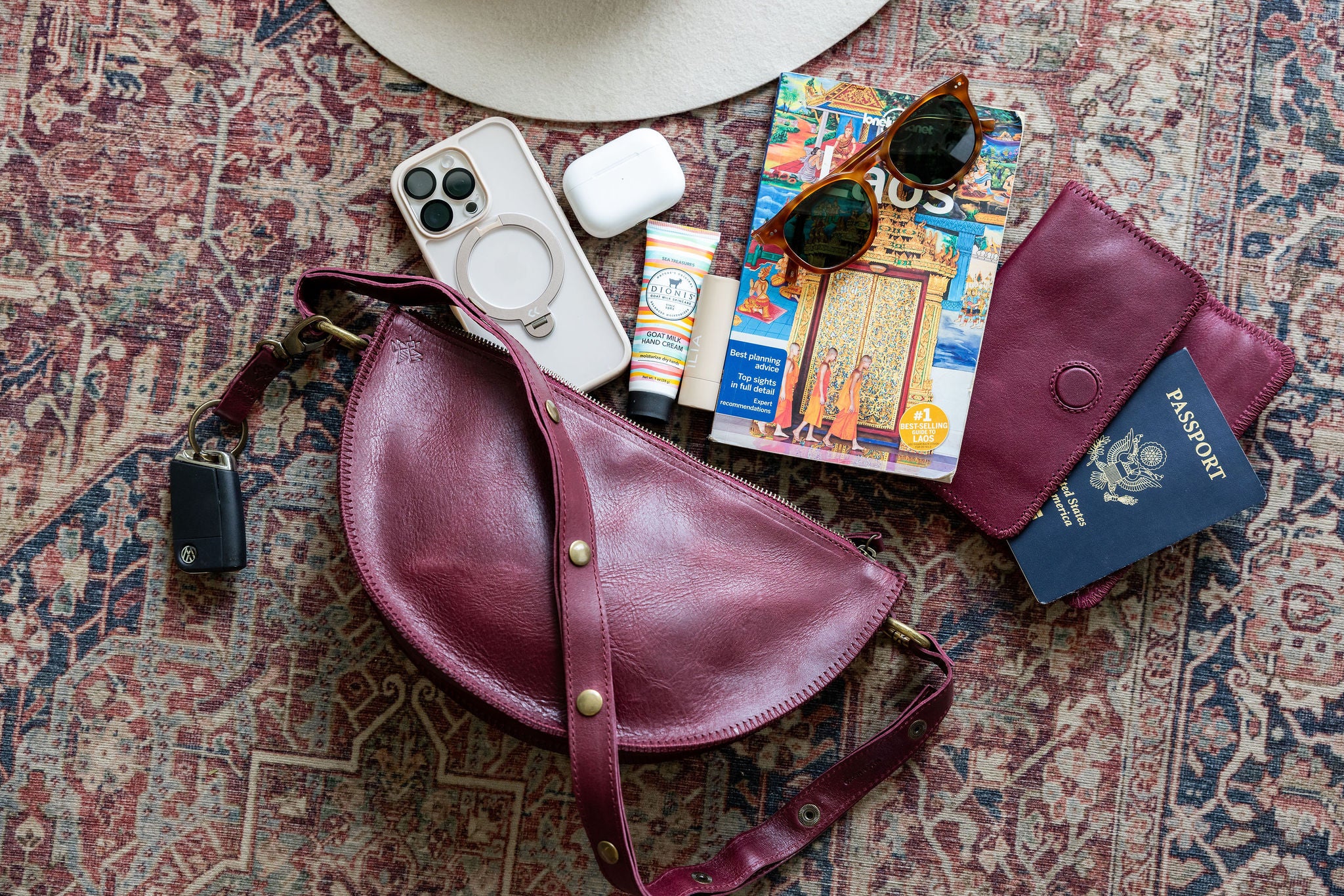 Cherry leather bag with travel items including a phone, sunglasses, passport, and travel guide on a patterned surface.