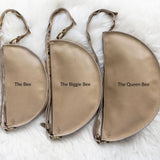 Three beige pouches labeled 'The Bee', 'The Biggie Bee', and 'The Queen Bee' on a white fur background.