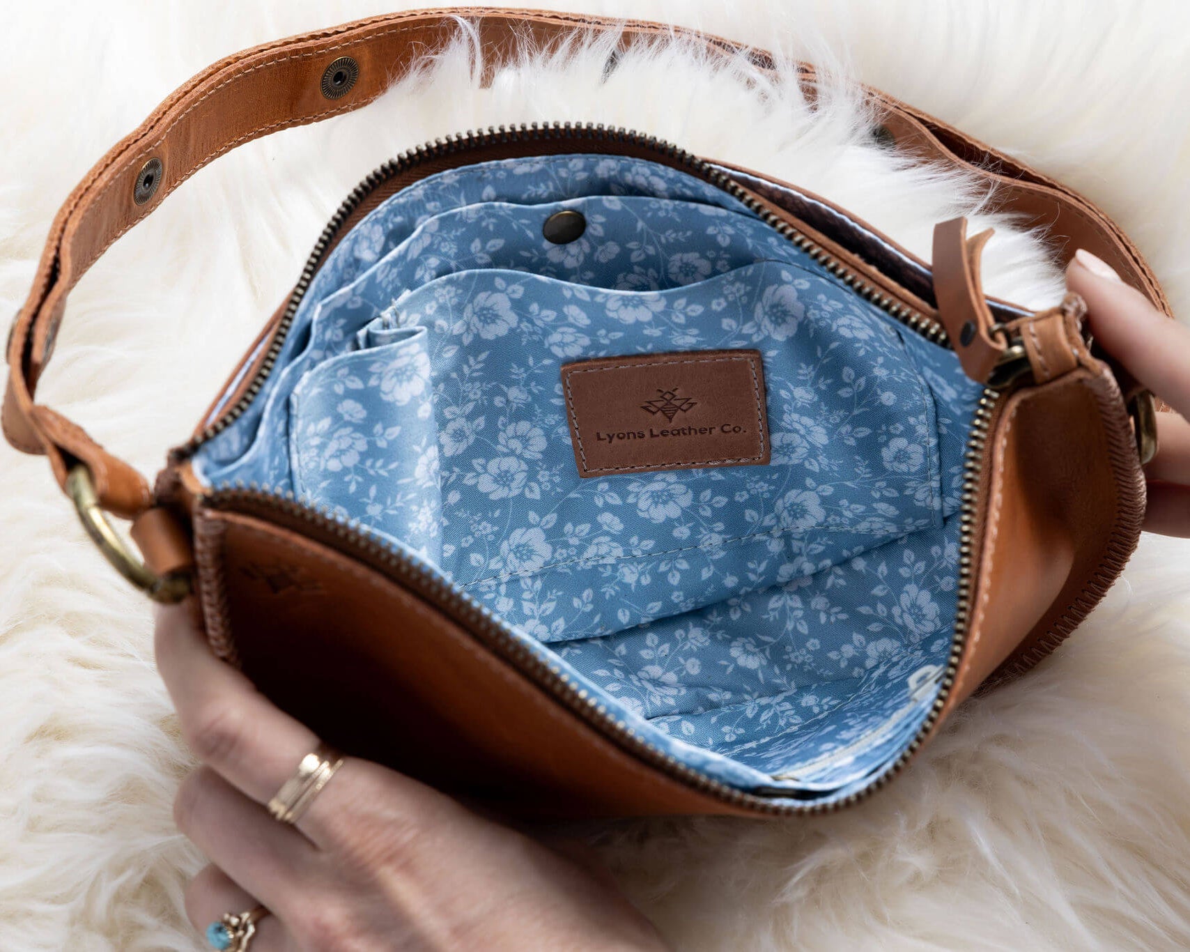 Brown leather bag with blue interior and floral pattern, held by a hand on a white fluffy surface.