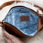 Brown leather bag with blue interior and floral pattern, held by a hand on a white fluffy surface.