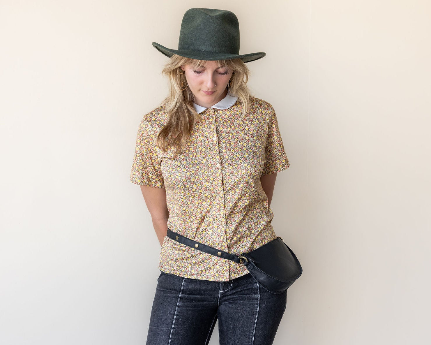 Woman wearing a patterned shirt, jeans, and a hat against a plain background