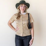 Woman wearing a patterned shirt, dark jeans, and a hat against a plain background