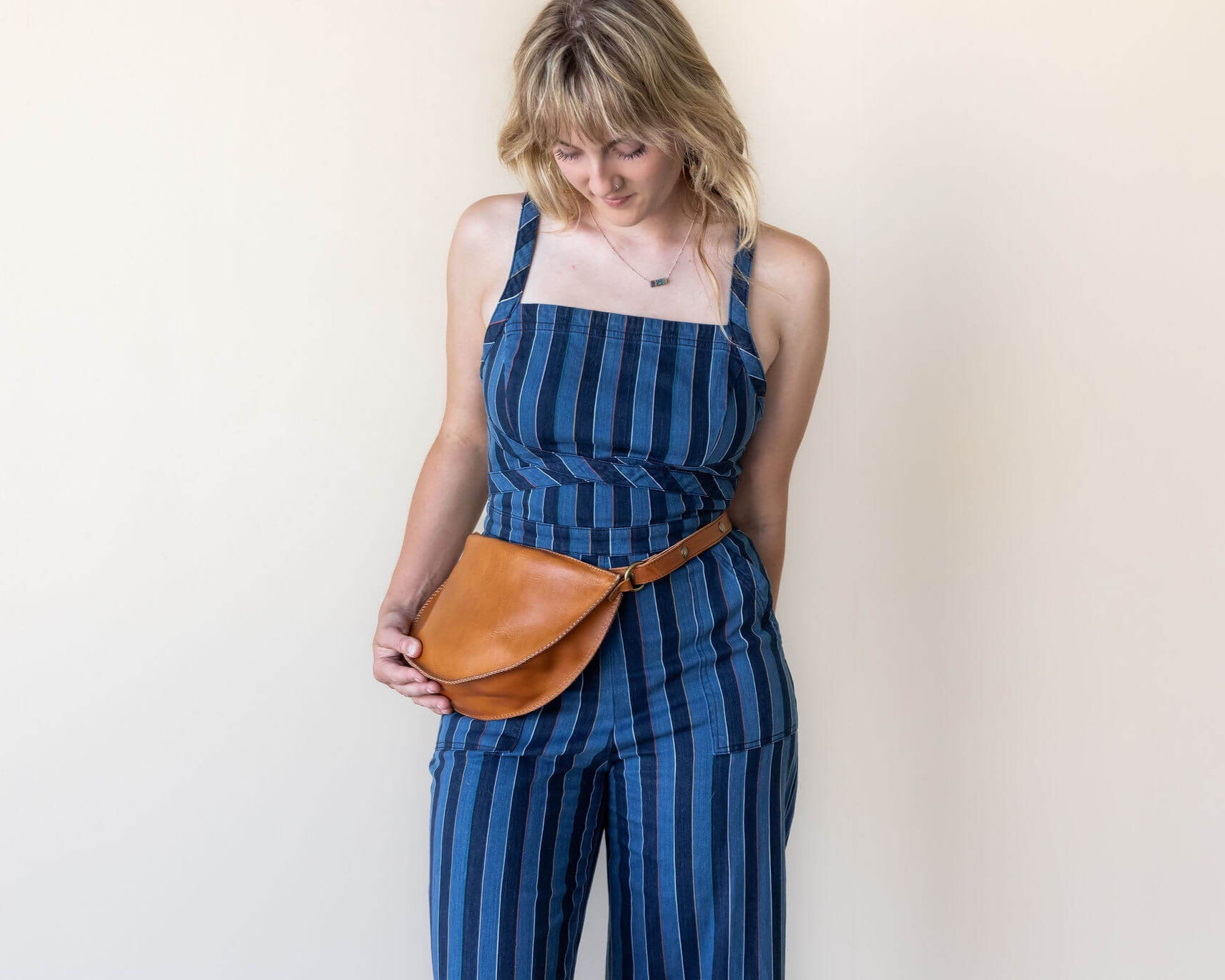 Woman wearing a blue striped jumpsuit with a brown leather belt bag against a plain background