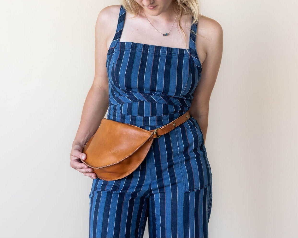 Woman wearing a blue striped jumpsuit with a brown leather belt bag against a plain background
