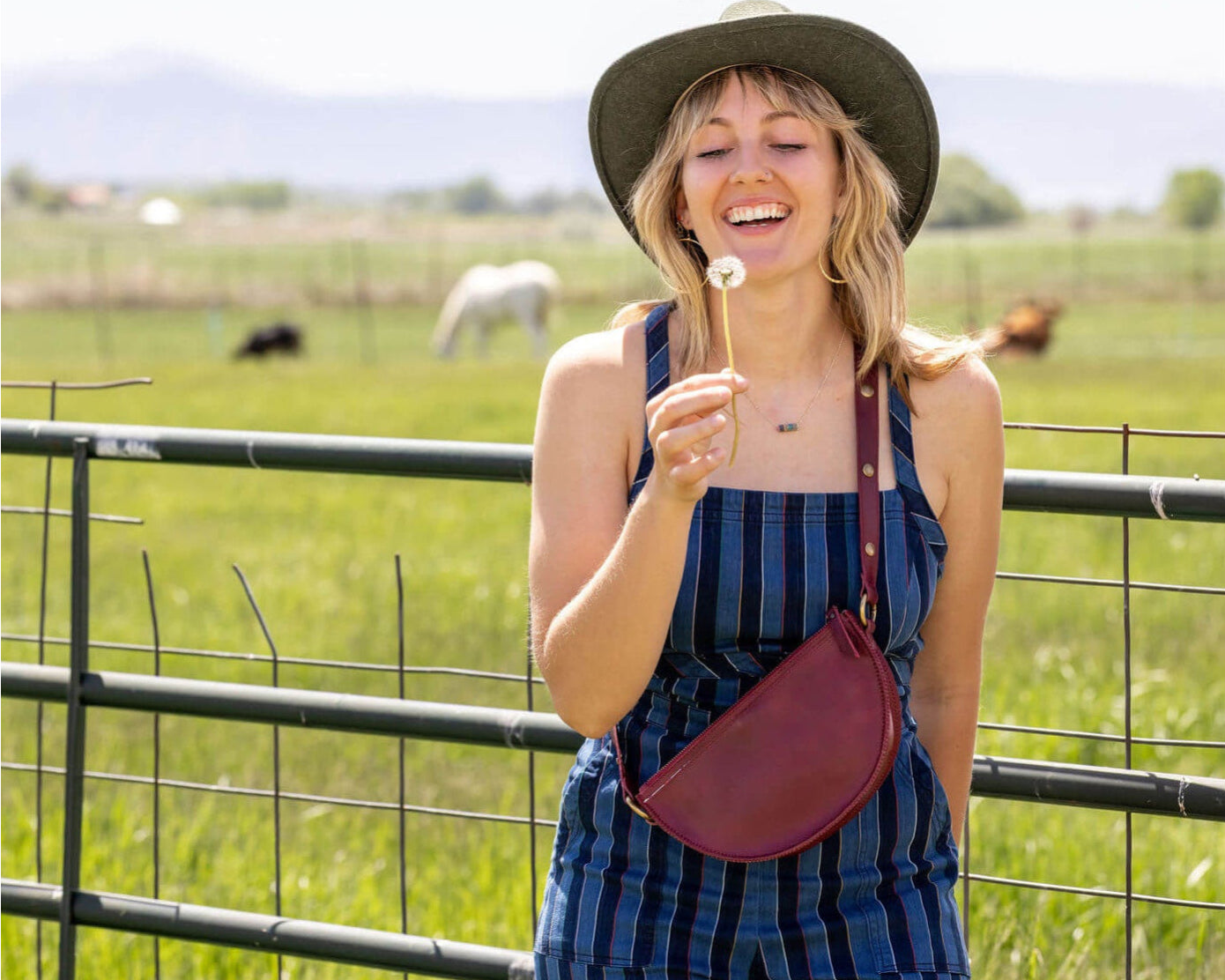 Woman in a striped dress and hat holding a dandelion in a field with animals in the background