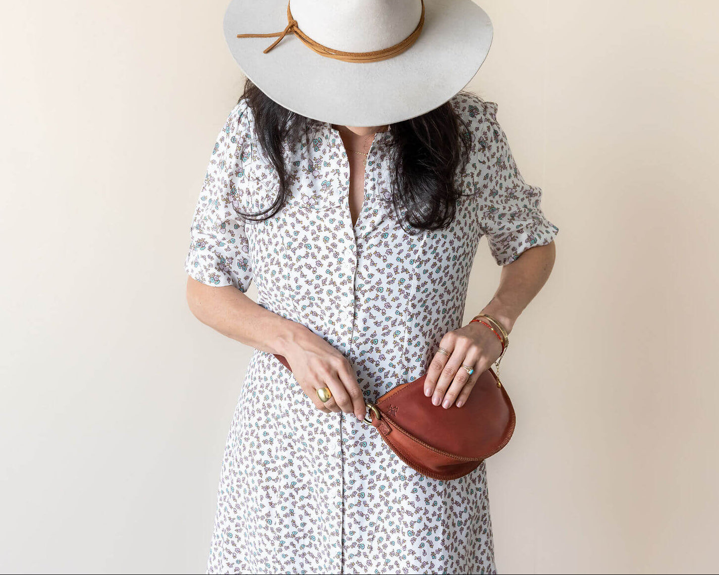 Woman wearing a white dress with black patterns and a wide-brimmed hat, holding a brown leather handbag against a beige background.