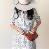 Woman wearing a white dress with black patterns and a wide-brimmed hat, holding a brown leather handbag against a beige background.