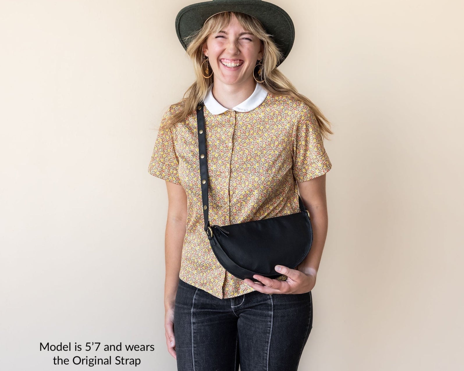 Woman wearing a gold patterned shirt, black hat, and carrying a black bag against a beige background.