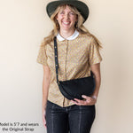 Woman wearing a gold patterned shirt, black hat, and carrying a black bag against a beige background.