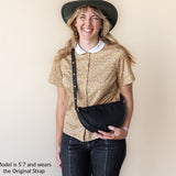 Woman wearing a gold patterned shirt, black hat, and carrying a black bag against a beige background.