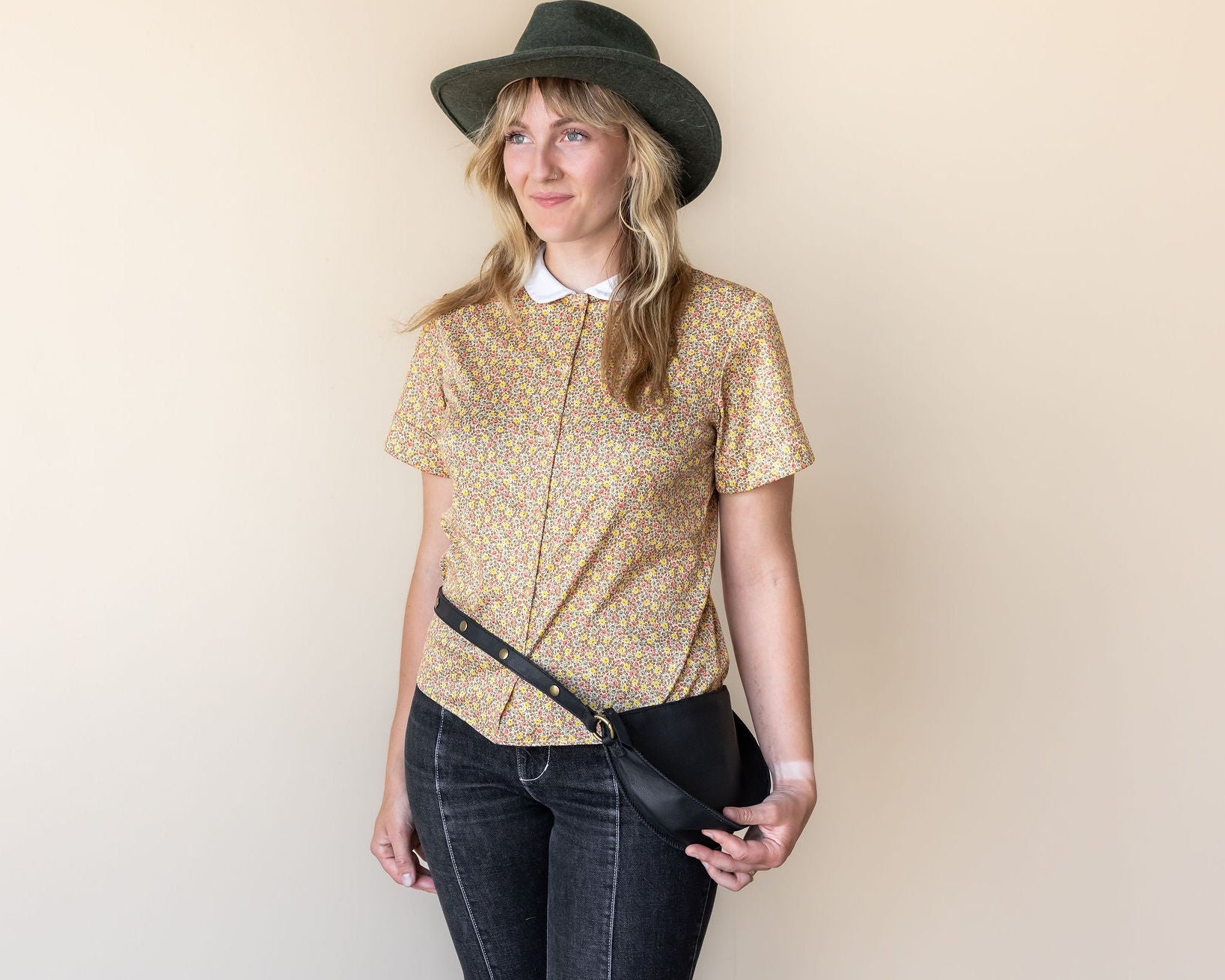 Woman wearing a patterned blouse, jeans, and a hat against a plain background
