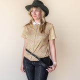 Woman wearing a patterned blouse, jeans, and a hat against a plain background