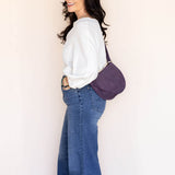 Woman wearing a white shirt and blue jeans with a purple bag against a plain background