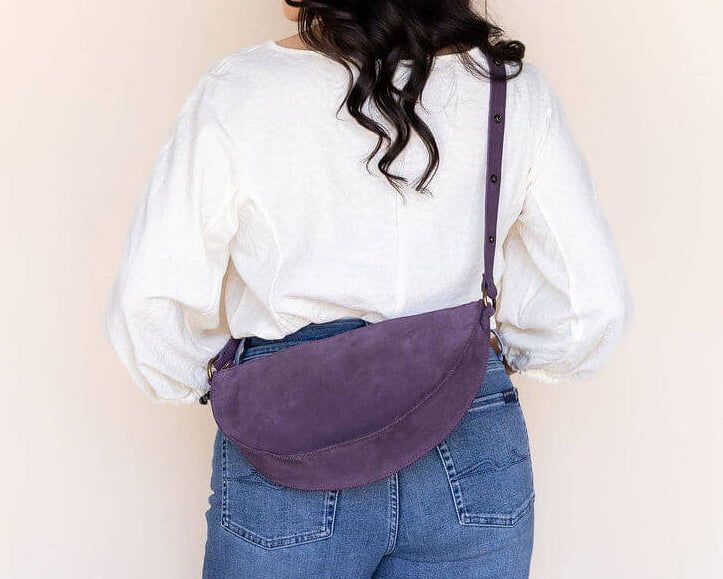 Person wearing a white shirt and blue jeans with a purple bag over their shoulder against a plain background