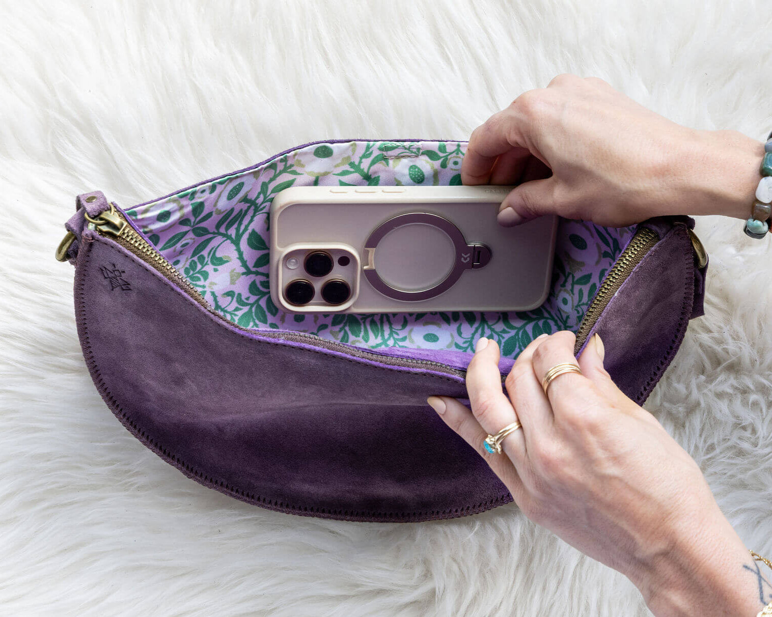 Purple pouch with floral interior containing a phone case, held by hands on a white fluffy surface.
