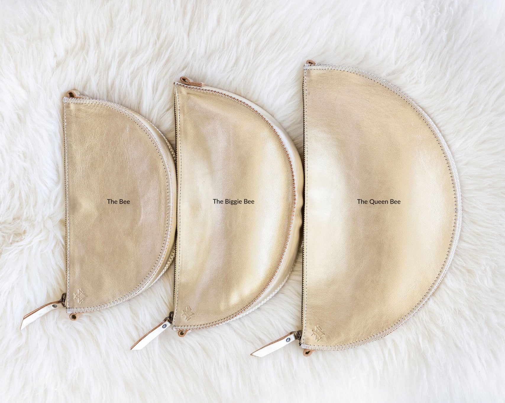 Three beige pouches labeled 'The Bee', 'The Bigger Bee', and 'The Queen Bee' on a white fur background.