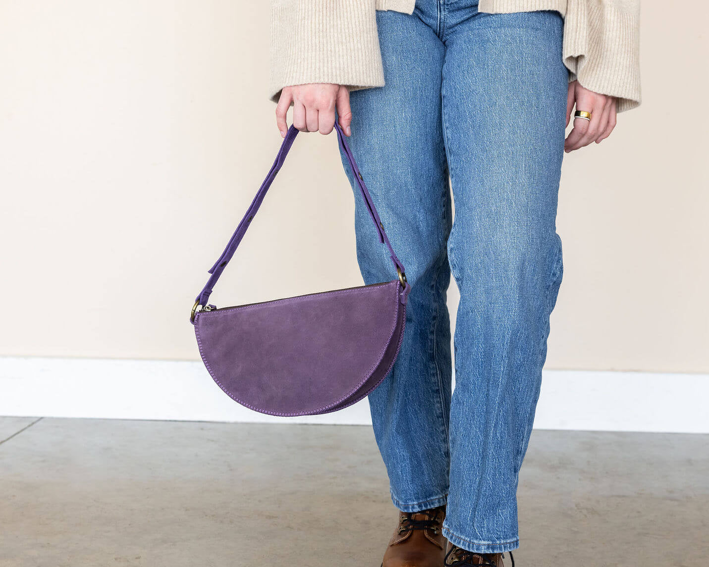 Person holding a purple handbag wearing blue jeans and brown shoes on a plain background