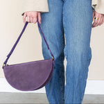 Person holding a purple handbag wearing blue jeans and brown shoes on a plain background