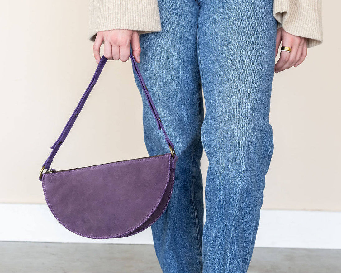 Person holding a purple handbag wearing blue jeans and brown shoes on a plain background
