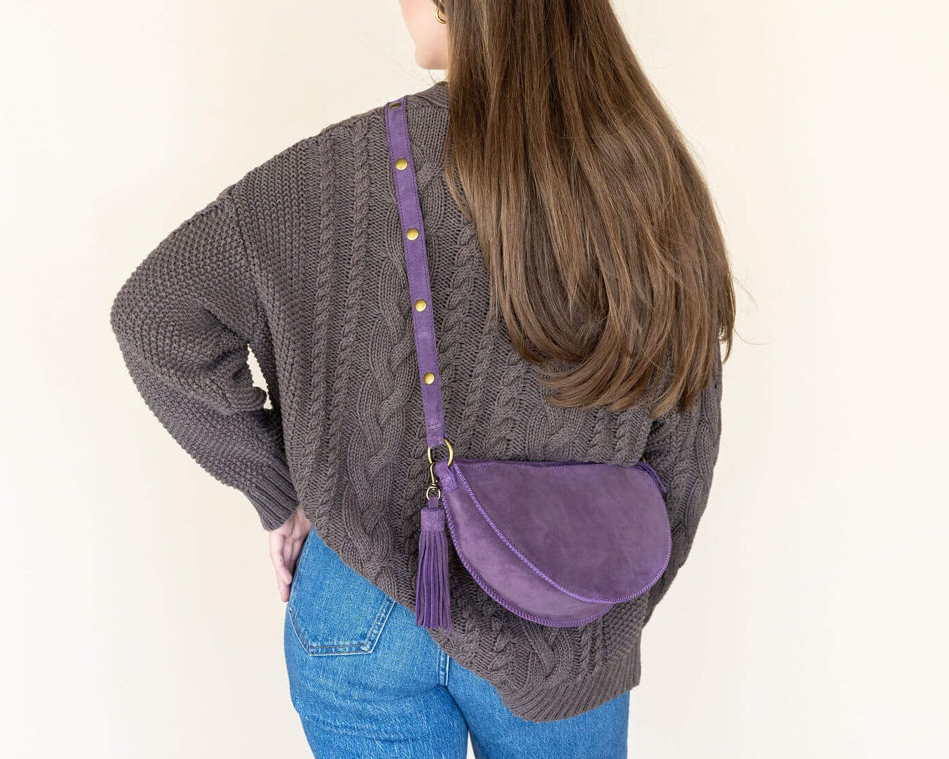 Person wearing a gray sweater and blue jeans with a purple crossbody bag on a plain background