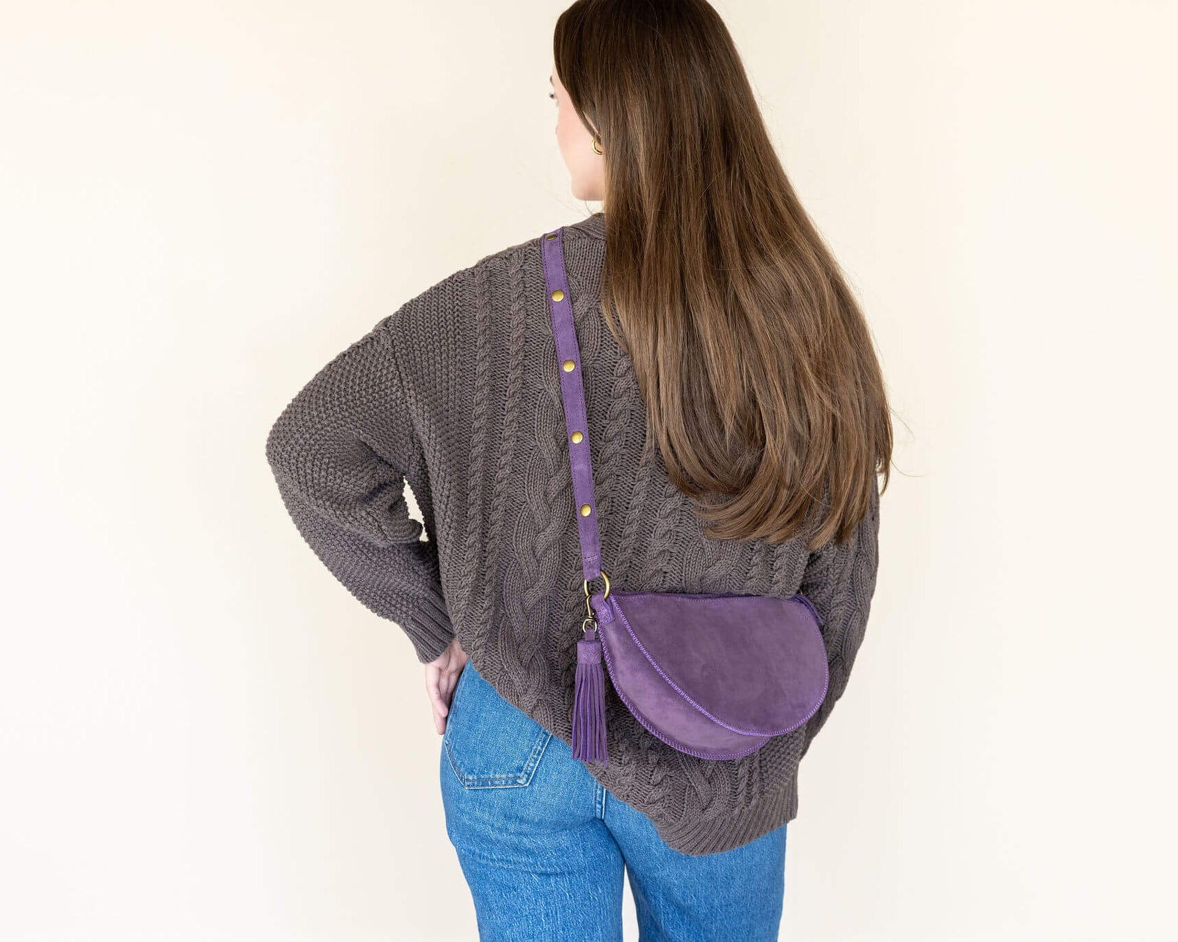 Person wearing a gray sweater and blue jeans with a purple crossbody bag on a plain background