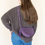 Person wearing a gray sweater and blue jeans with a purple crossbody bag on a plain background