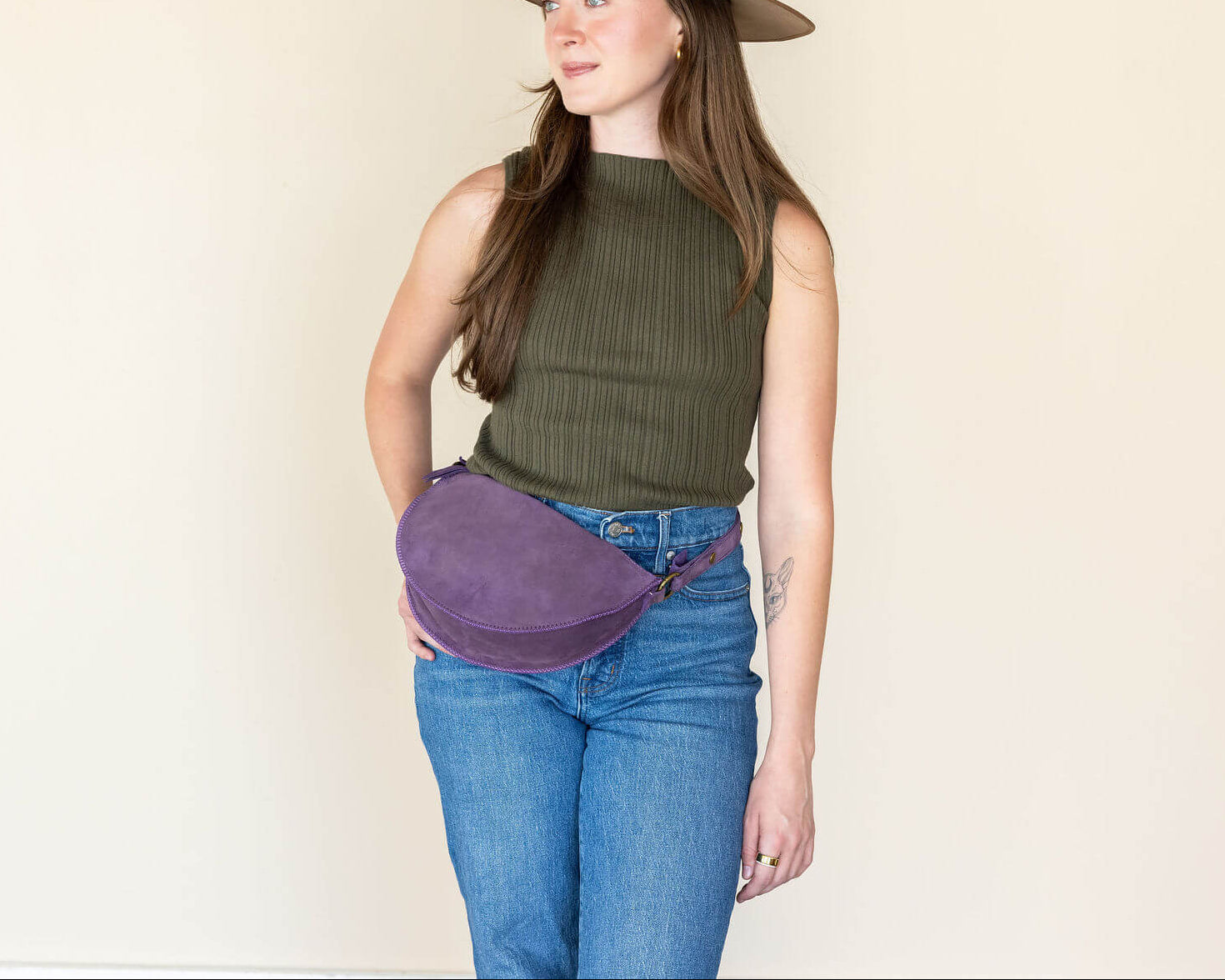 Woman wearing a brown hat, green sleeveless top, blue jeans, and a purple fanny pack against a plain background