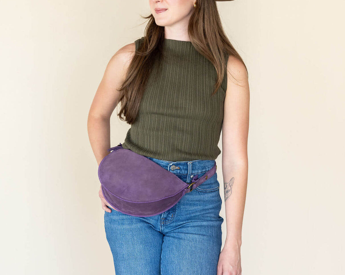 Woman wearing a brown hat, green sleeveless top, blue jeans, and a purple fanny pack against a plain background