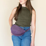 Woman wearing a brown hat, green sleeveless top, blue jeans, and a purple fanny pack against a plain background